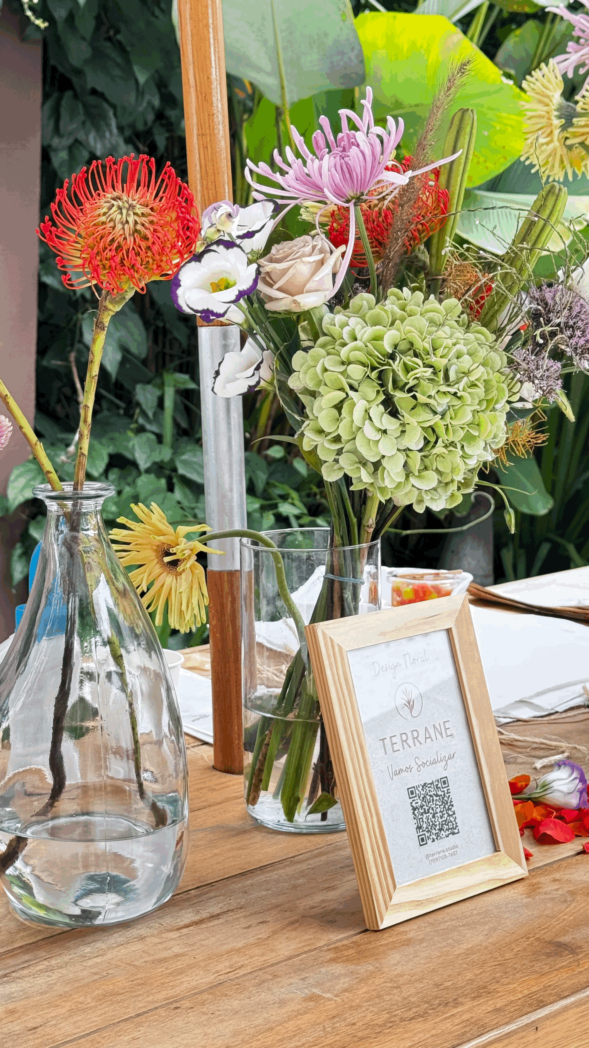 Arranjos florais TERRANE em vasos de vidro sobre mesa de madeira, com placa de identificação da marca em ambiente de jardim
